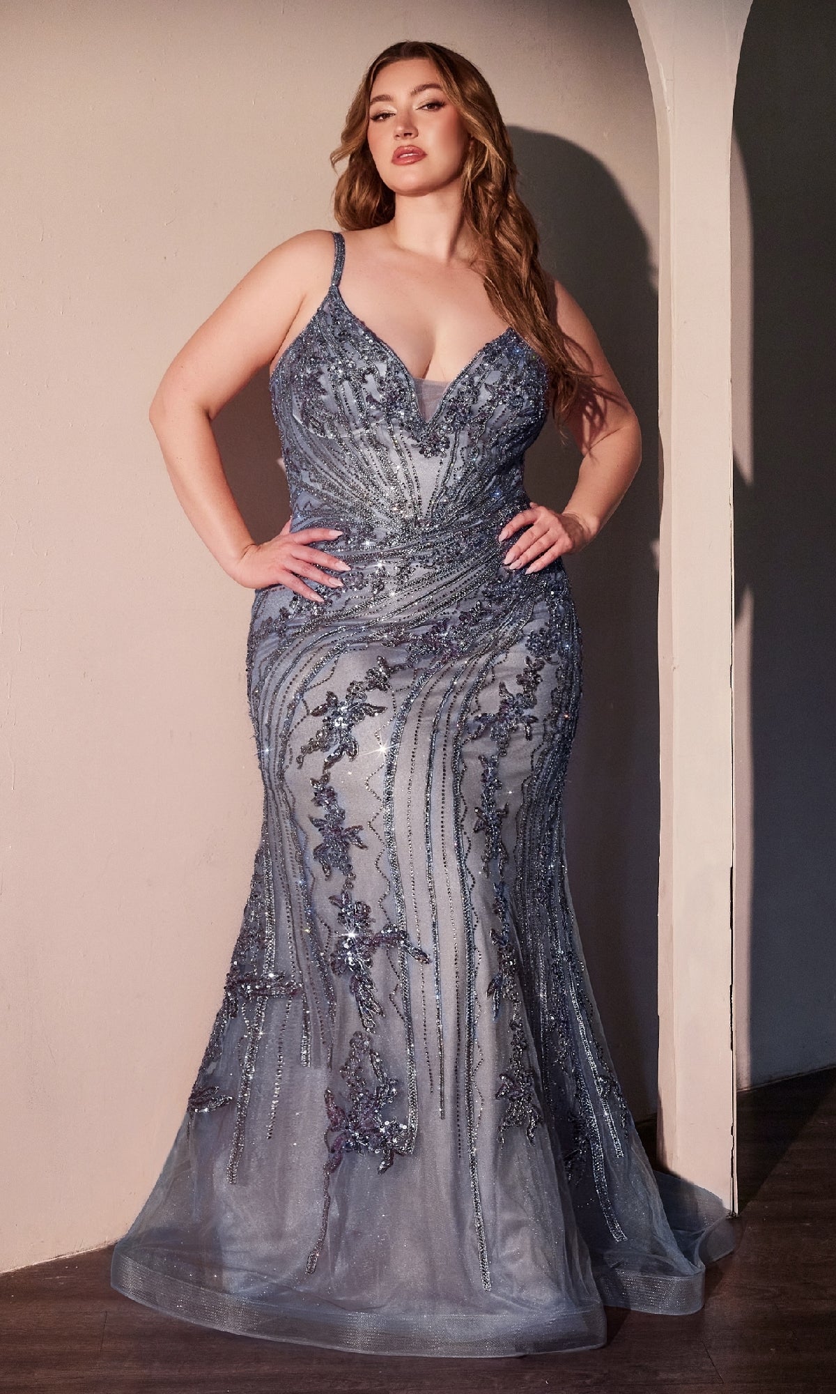 A woman stands indoors wearing the Plus-Size Long Beaded Prom Dress: Ladivine CC0389C, a fitted blue floor-length gown with sparkling sequin and lace details, her hands elegantly resting on her hips.